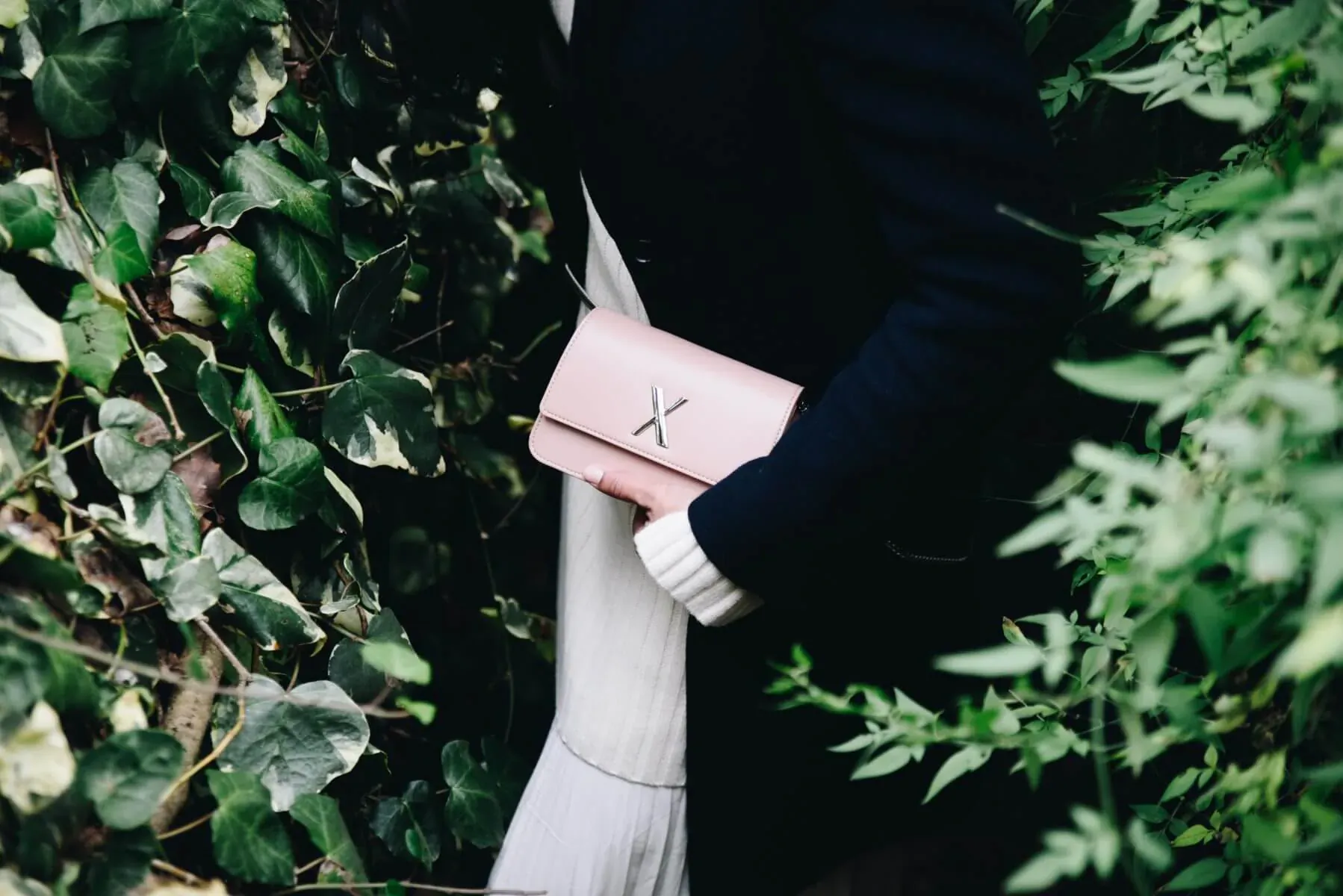 Person in black coat and white skirt holding pink 'X' clutch purse amid lush green foliage
