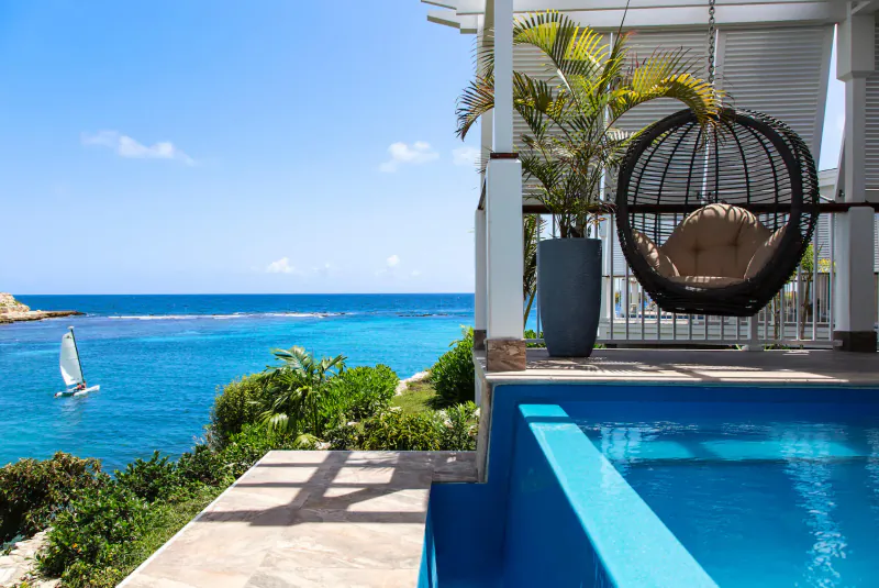 Luxury villa balcony at Hammock Cove, Antigua: egg chair, infinity pool overlooking turquoise sea with sailboat.