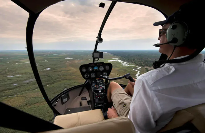 Helicopter pilot in headset flying over African bush landscape with river and wetlands visible through cockpit.