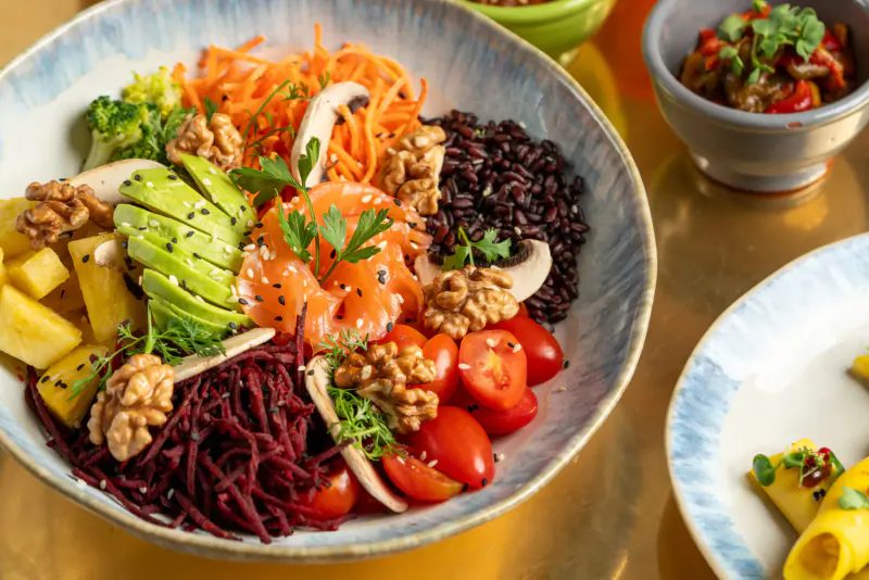 Vibrant poke bowl with salmon, black rice, purple noodles, avocado, pineapple, walnuts, mushrooms, and tomatoes in blue bowl
