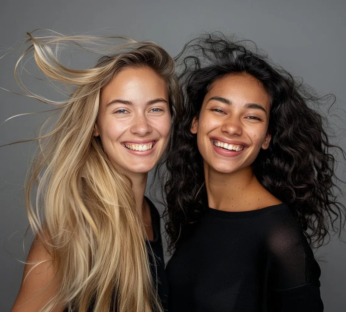 Two smiling women with flowing blonde and curly black hair, posing together against gray background