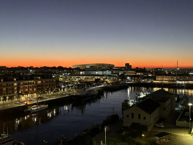 Twilight aerial view of Cape Town's V&A Waterfront stadium, marina, and riverside buildings at sunset.