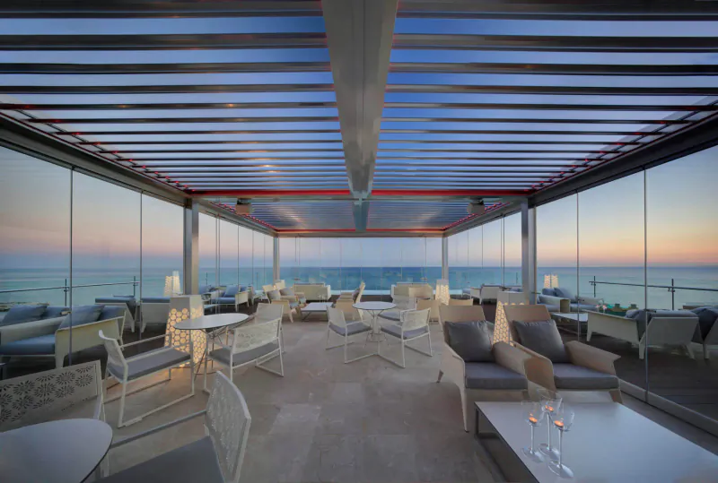 Amàre Marbella rooftop lounge with white chairs and tables under blue pergola at sunset over sea.