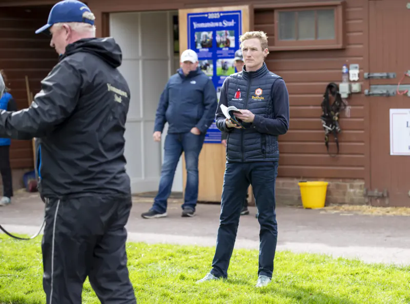 At Tattersalls, Emma Blunt in maroon vest holds papers outside barn with horseshoe and yellow bucket; men in dark jackets nearby.