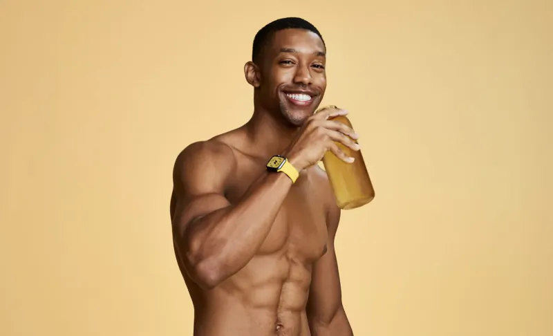 Muscular Black man shirtless smiling, holding yellow bottle with yellow wristband, yellow background