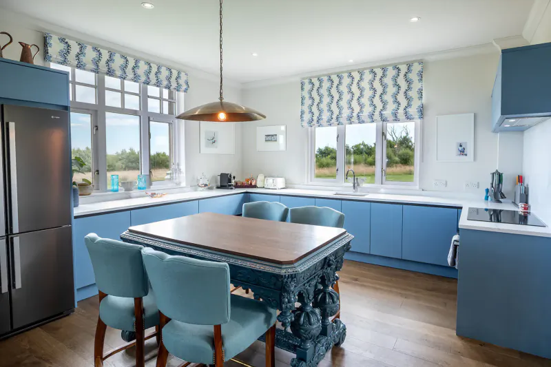 Modern luxury kitchen with blue cabinets, ornate wooden dining table, chairs, and windows overlooking countryside.