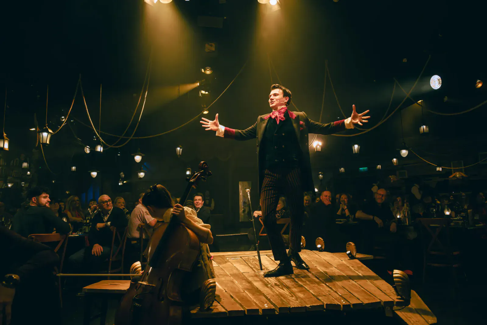 Man in black suit with red ascot dramatically poses arms outstretched on wooden stage, lit by lanterns in immersive theatre show.