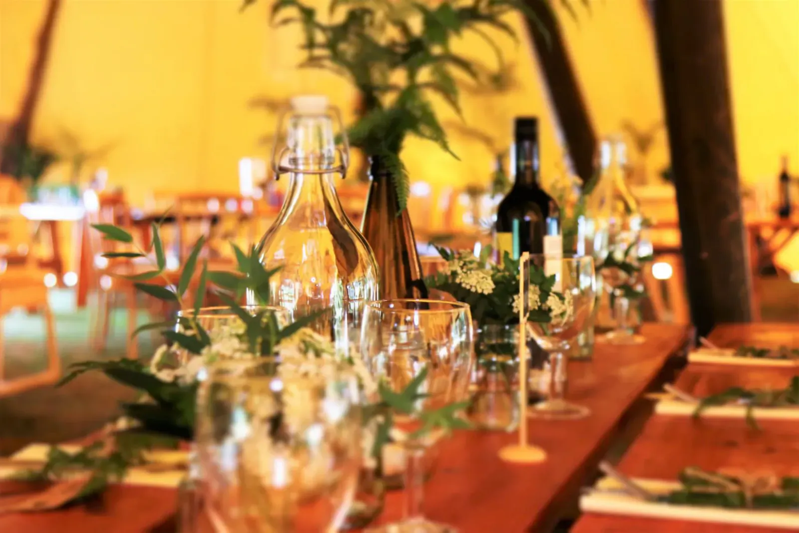 Close-up of Wahaca supper club table in tent: wooden table with wine bottles, glasses, white flowers, greenery, and plates.
