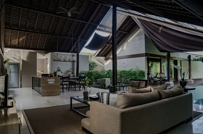 Open-air luxury villa living room in Bali with modern kitchen, beige sofa, plants, and infinity pool.