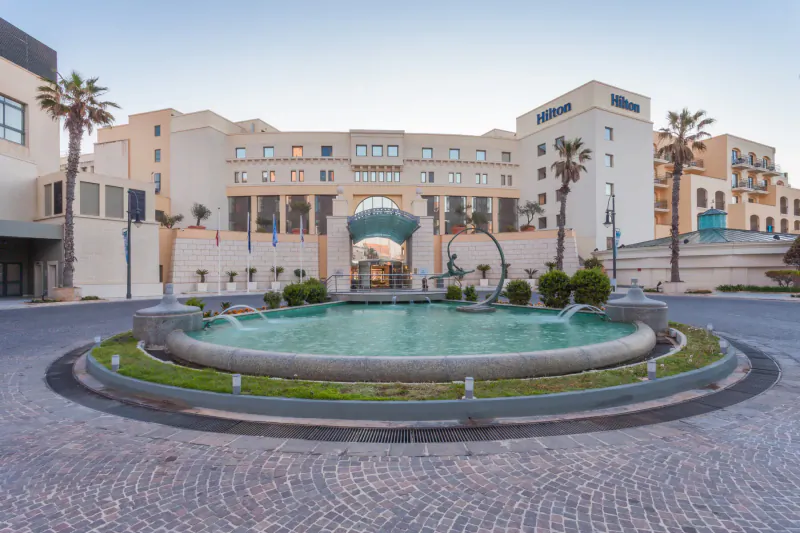Hilton Malta hotel facade with central fountain, palm trees, and landscaped gardens in circular driveway