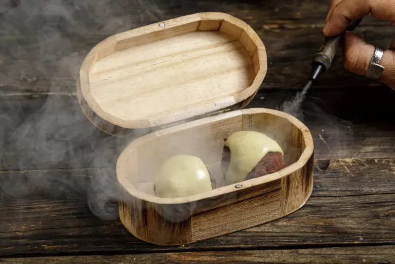 Open wooden box on rustic table holding two cheese-filled spheres with steam rising, hand using blowtorch.