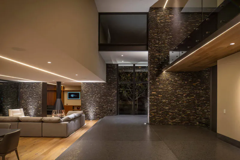 Modern Mexican living room with brick walls, wooden stairs, gray sofa, TV, and large windows