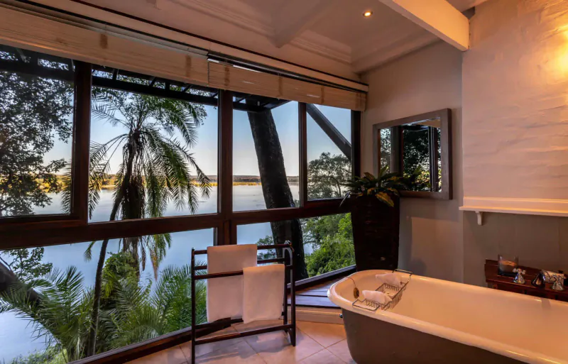 Luxurious bathroom at The River Club with freestanding tub, towels, and panoramic view of palm trees and river.