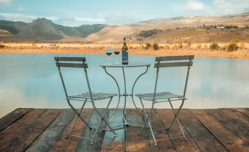 Two chairs and wine bottle with glasses on table by lake at South Hill Vineyards, mountain backdrop.