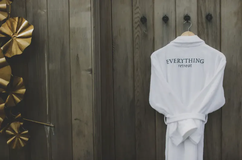 White bathrobe with 'Everything Retreat' embroidered on back, hanging on wooden door hook beside gold fan decor.