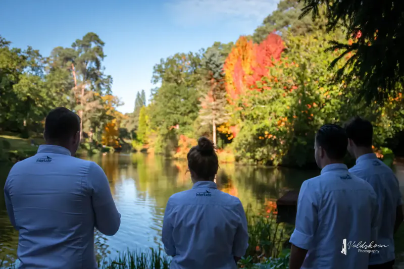 Three people in white shirts stand backs to camera, viewing serene autumn lake with red foliage and trees.