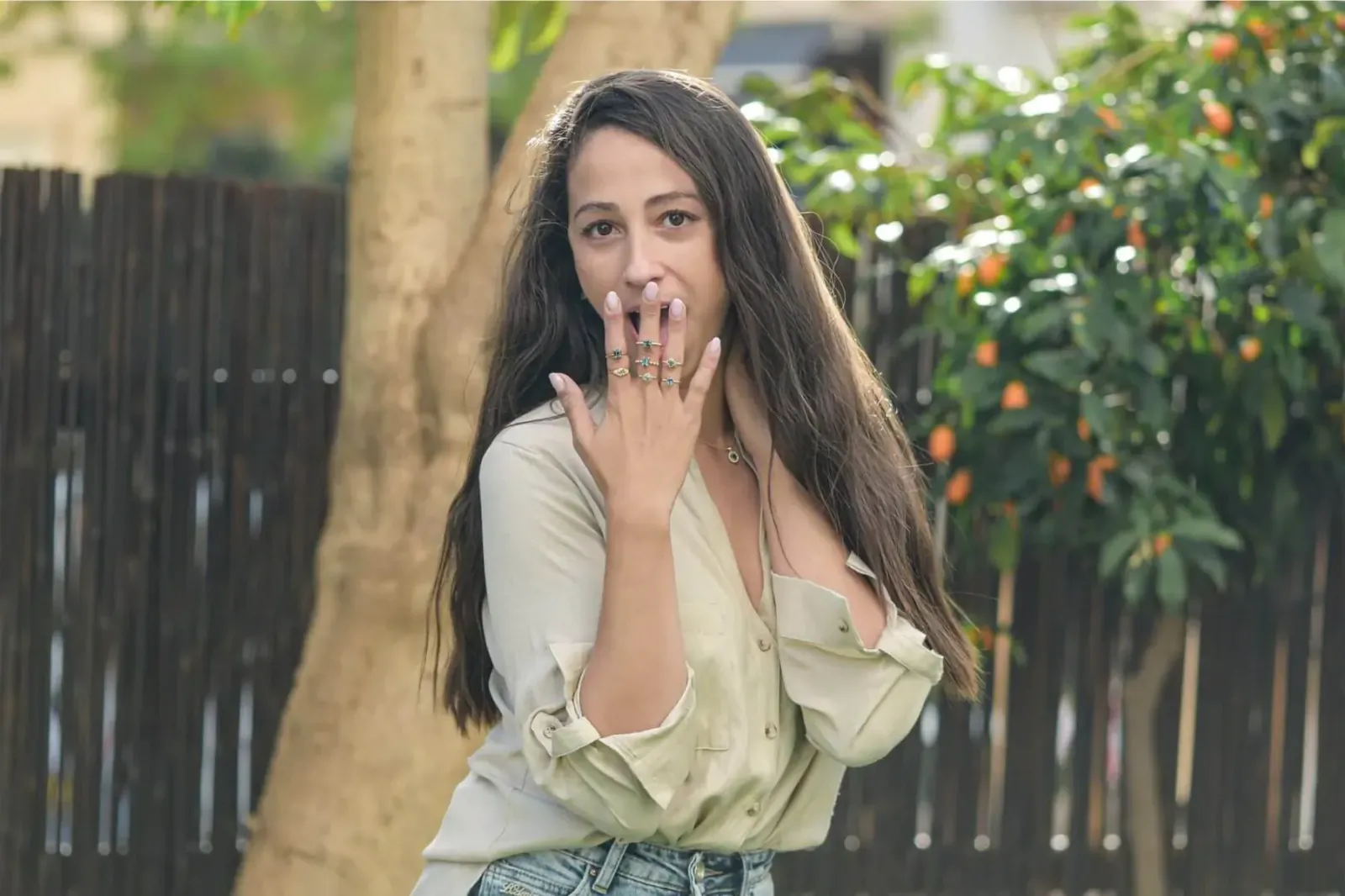 Woman with surprised expression, hands near mouth showing rings, in beige blouse and jeans by tree with oranges, Mogga Jewels featured