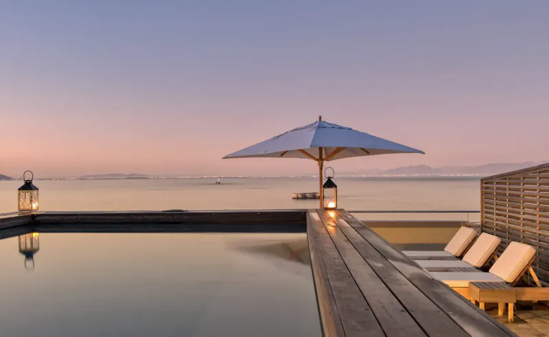 Twilight infinity pool at Tintswalo Boulders Boutique Hotel overlooking Simon's Town sea, with loungers, umbrella, and lanterns.