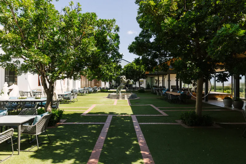 Sunny courtyard at Benguela Cove wine estate with fountain, trees, grass paths, and outdoor tables