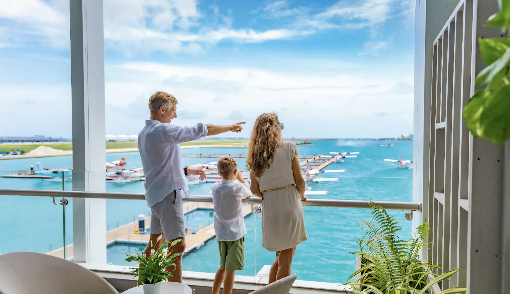 Family on balcony at Lily Hotels overlooking Noovilu Seaplane Terminal marina and water, father pointing out view.