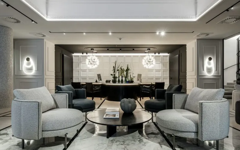 Elegant modern hotel lobby in Vienna with curved gray sofas, black round coffee table with vase and magazine, pendant lights, plants.