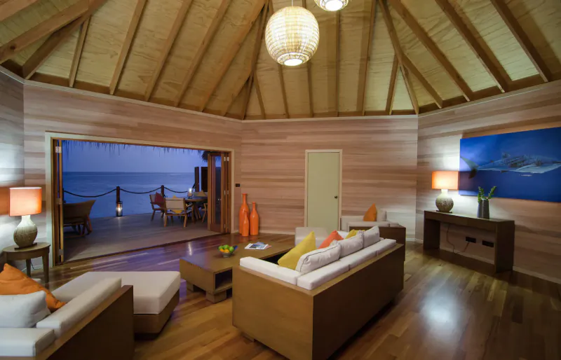 Spacious thatched-roof resort living room with wooden furniture, orange cushions, lamps, and ocean deck view at sunset.