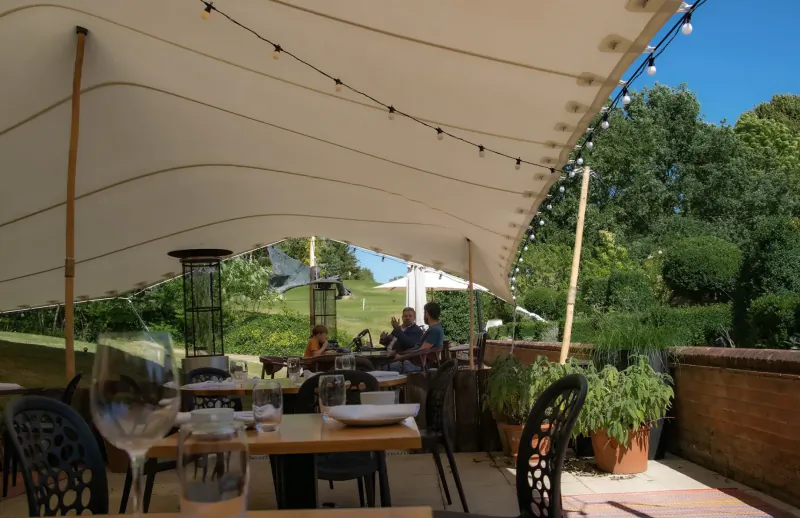 Outdoor terrace dining under beige tent with string lights, patrons at wooden tables, garden and golf course backdrop at Donnington Valley.