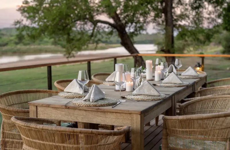 Elegantly set wooden dining table with white napkins and candles on outdoor deck overlooking river and trees at sunset.