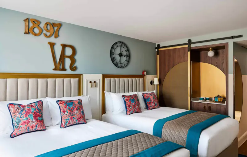Hotel Indigo Chester room with two beds, pink floral pillows, '1897 VR' wall sign, clock, and barn door.