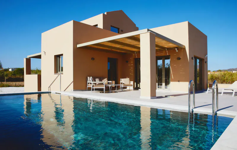 Beige modern villa with wooden pergola, outdoor seating, and infinity pool reflecting blue sky at Cavo Spada resort, Crete.