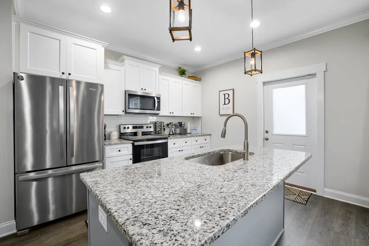 Modern white kitchen with stainless steel fridge, granite island, pendant lights, and 'B' wall sign