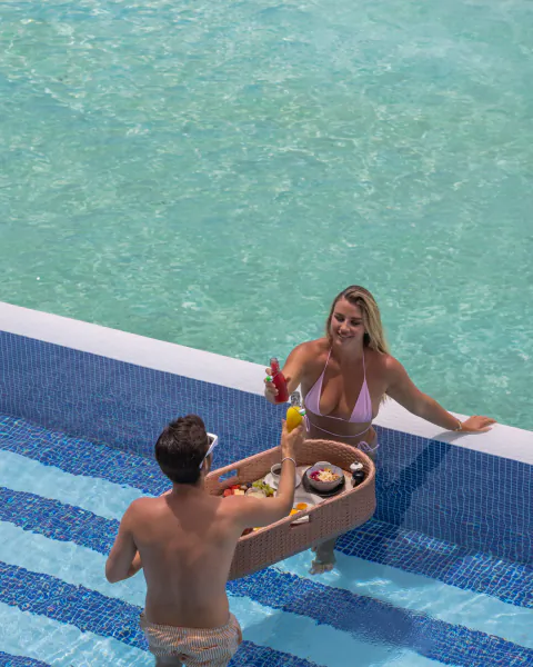 Man and woman in pool at Maldives resort, man holding wooden tray of sushi and drinks toward her.