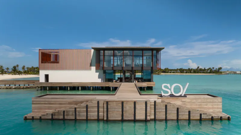 Modern 'SO/' overwater building on turquoise lagoon at Crossroads Maldives, with wooden pier and beach backdrop.