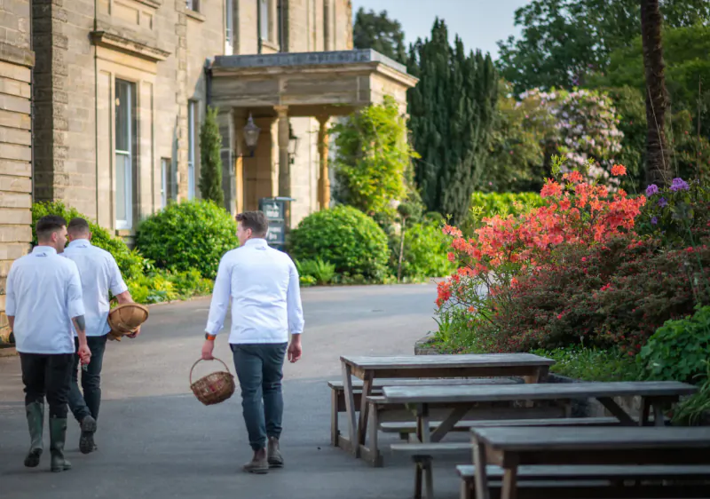 Three chefs in white uniforms carrying wicker baskets walk past picnic tables toward a stone manor house amid gardens and azaleas.