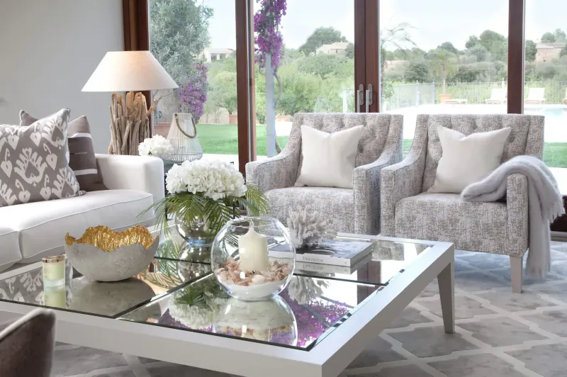 Elegant living room with white sofa, tufted armchairs, glass coffee table with flowers, candles, and garden view through large windows.