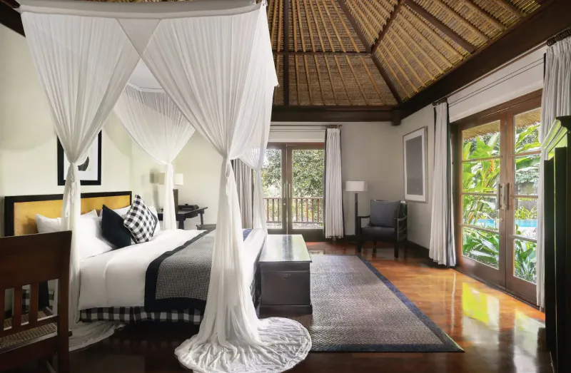 Luxurious Balinese villa bedroom with white canopy bed, wooden ceiling, and sliding doors to lush garden overlooking Ayung River.