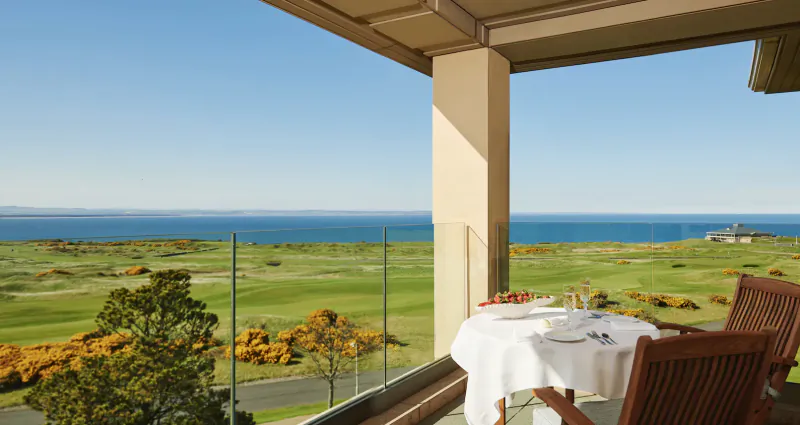 Luxury balcony dining setup with white tablecloth, wine glasses, overlooking Scottish golf course and sea.