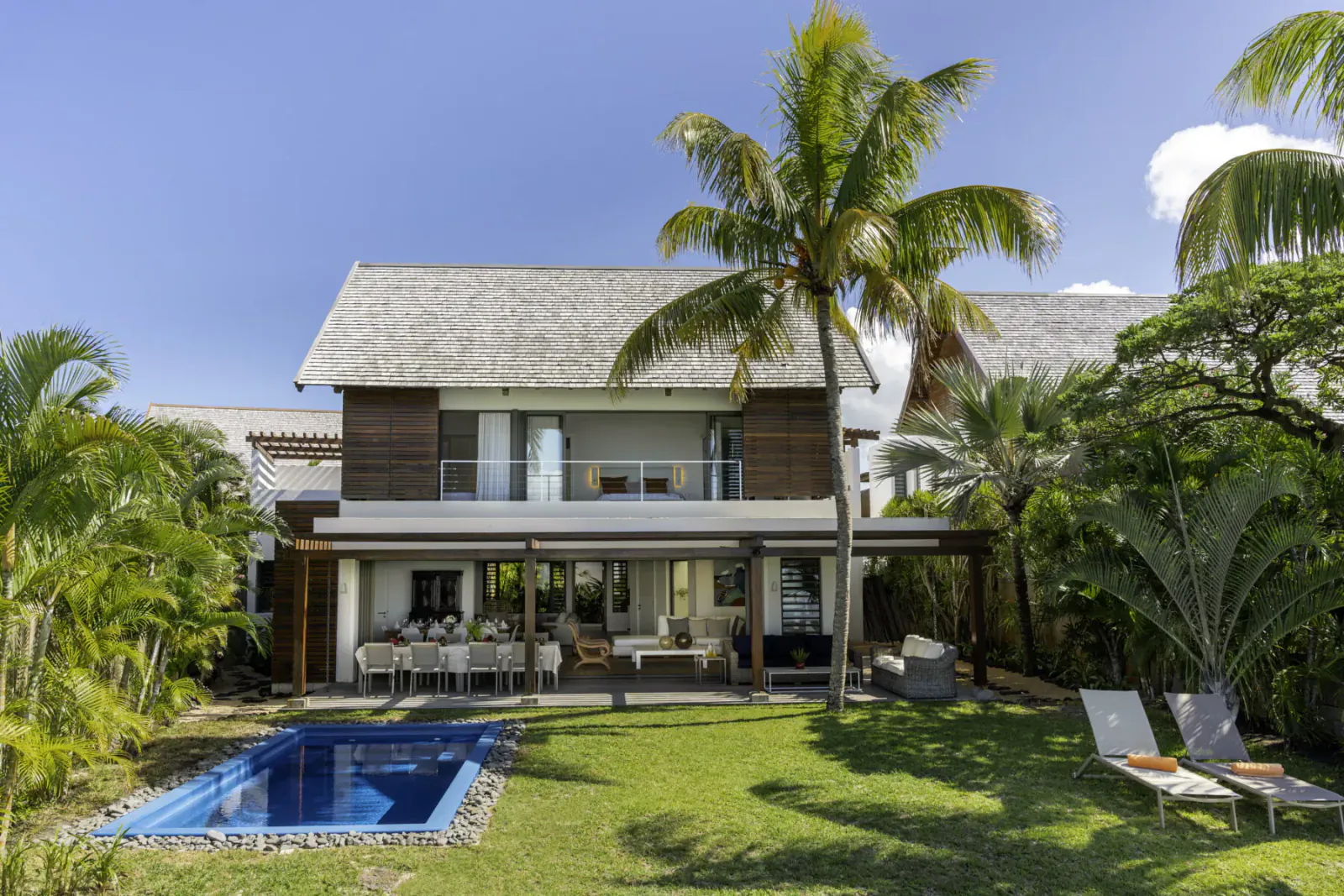 Modern thatched-roof villa in Mauritius with infinity pool, outdoor dining, lounge chairs, and palm trees under blue sky.
