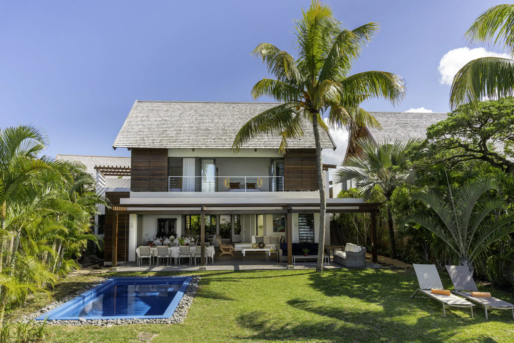 Modern thatched-roof villa in Mauritius with infinity pool, outdoor dining, lounge chairs, and palm trees under blue sky.