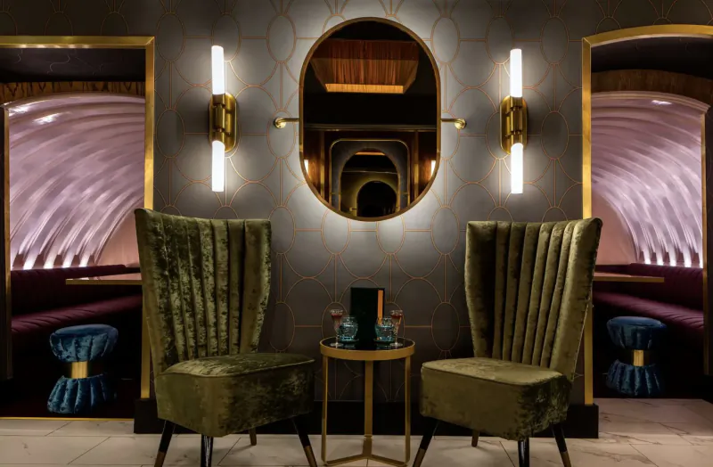 Elegant Disrepute Bar interior in Soho: two green velvet chairs at gold table with cocktails, arched mirrors, ambient lighting.