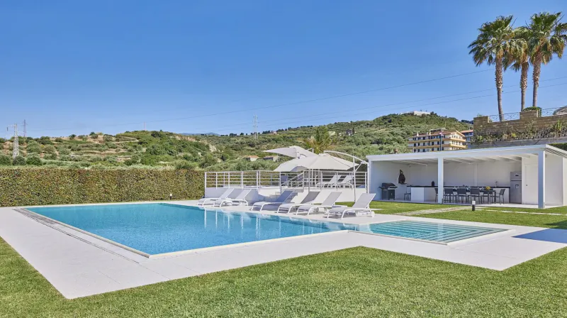 Luxury infinity pool with white loungers and bar, palm trees, hedges, and hills under blue sky at Wish Sicily resort.