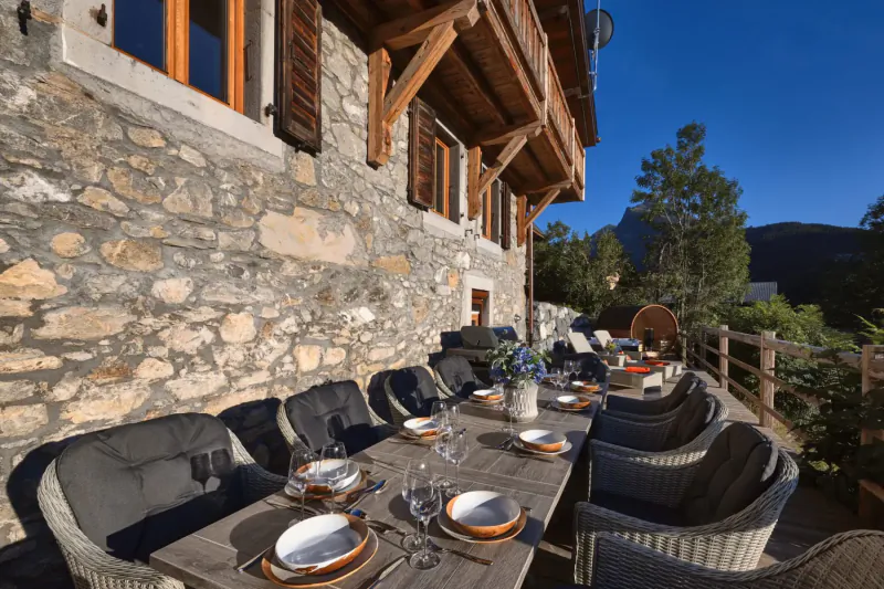 Elegant outdoor dining table set on wooden deck of stone Hunter Chalets, with plates, wine glasses, flowers, alpine mountains backdrop.
