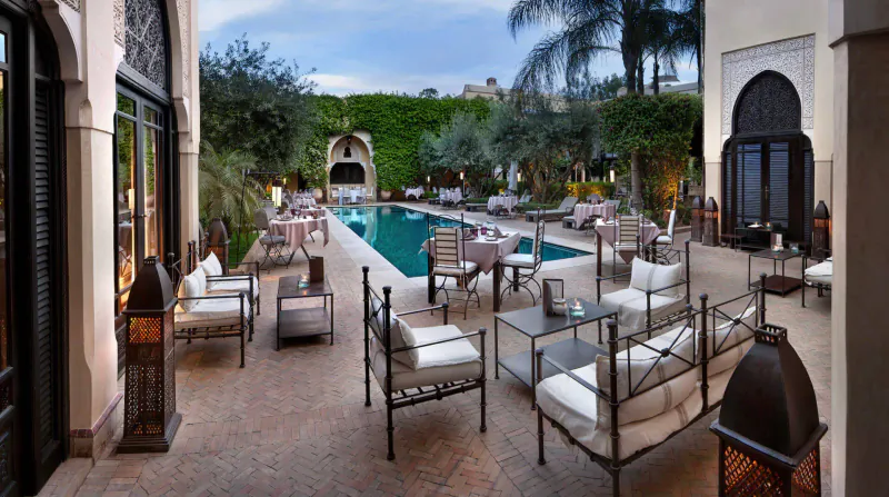 Luxurious Marrakech riad courtyard with infinity pool, white lounge chairs, lanterns, palm trees, and arched doorways at dusk.