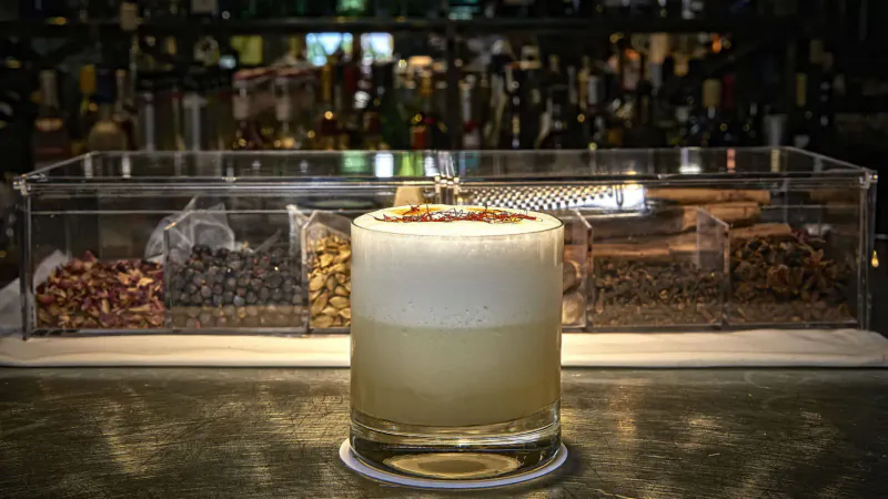 Winter cocktail with foamy top and yellow layer in glass, garnished, beside spices in acrylic tray on bar counter