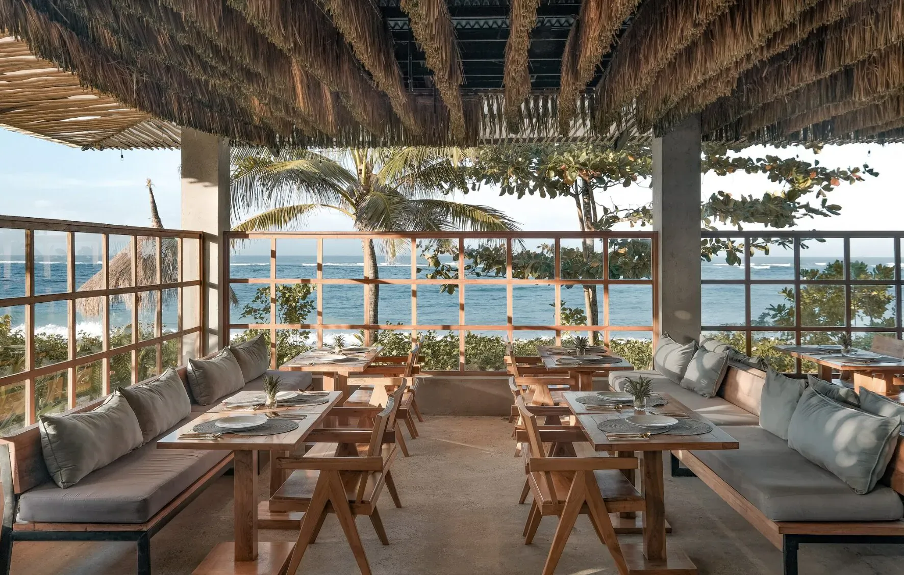Open-air beachside restaurant with wooden tables, cushioned benches, plants, and ocean views through bamboo screens.