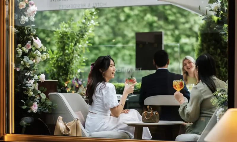 Group of four stylishly dressed people toasting with cocktails at a luxurious garden terrace table with flowers at The Athenaeum Hotel.