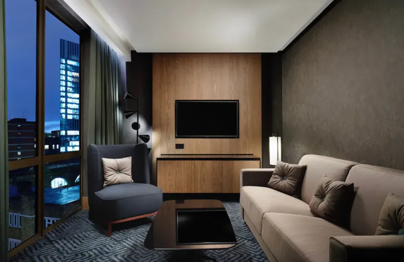 Modern hotel living room with L-shaped sofa, armchair, TV, lamps, and city skyline night view through windows.