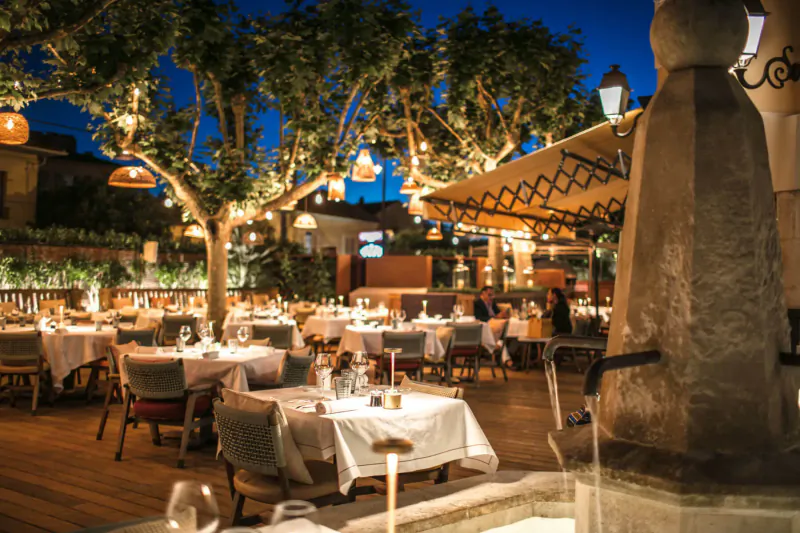 Elegant outdoor dining at Hôtel Byblos St Tropez at night, candlelit tables under strung lights and trees by stone fountain