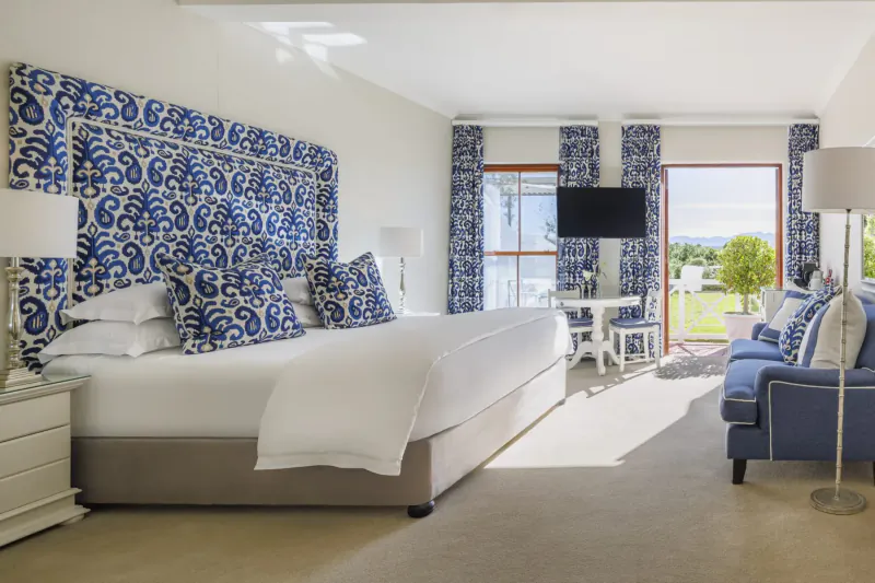 Elegant hotel bedroom with blue patterned headboard, white bed, blue sofa, TV, and garden view through windows.