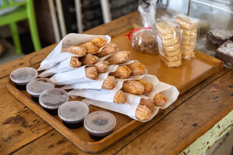 Assortment of churros in paper cones, cookies, and cups of sauce on wooden tray at Olympia Café